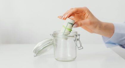 A close-up of a woman's hand putting rolled US dollar bills into a glass jar, symbolizing saving and budgeting.